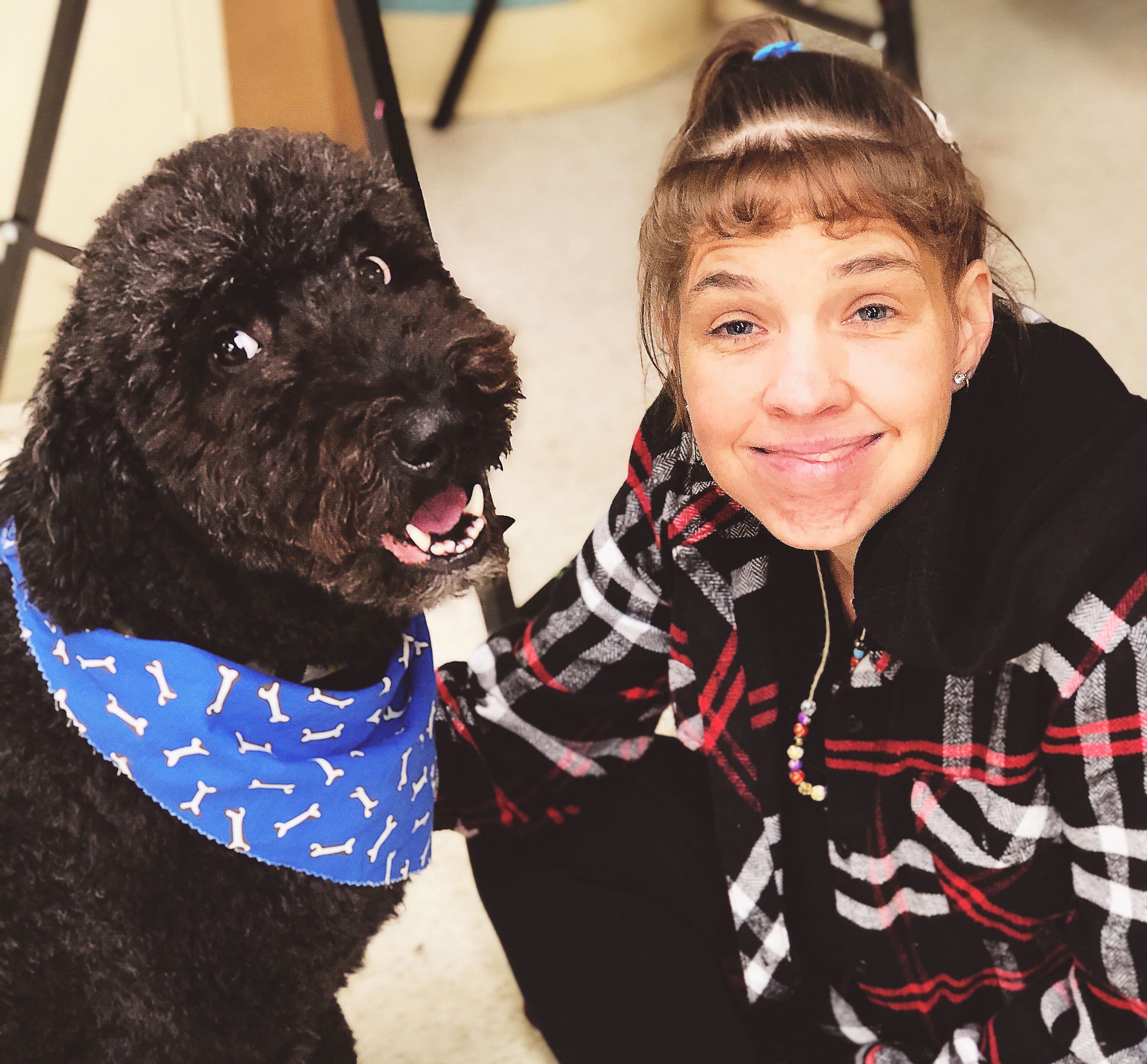 Female client smiling is kneeling down and face to face petting a happy and gentle looking black dog with a blue bandana that is part of the pet therapy program.