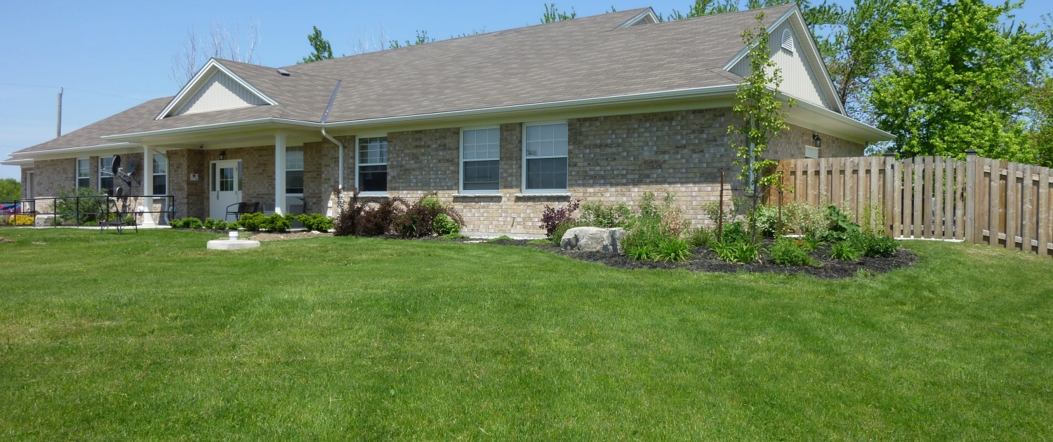 Large beige brick ranch-style bungalow has lots of front windows a covered entrance and a fenced area on the right side. It is surrounded by a well maintained lush green lawn and landscaped with trees and shrubs.