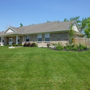 Large beige brick ranch-style bungalow has lots of front windows a covered entrance and a fenced area on the right side. It is surrounded by a well maintained lush green lawn and landscaped with trees and shrubs.