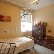 Client's bedroom with white bedspread and attached handrails for safety. 2 dark brown dressors of different sizes are up against the wall beside the window. The wall is decorated with a peices of art. A hoyer lift and belt is attached to the ceiling for transfer support.