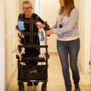 Female client using a walker is assisted by a female staff member while they walk together down a hall.