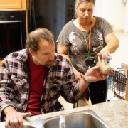 Male patient in a wheel chair sitting in front of a sink and female staff standing beside him are in the kitchen working together to put dishes in the dishwasher.