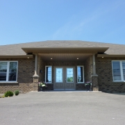 Brown brick bungalow with large double windowed entrance doors. A covered large entrance way is supported by brick and beige painted wooden pillars. A large paved driveway leads right up to the front of the house.