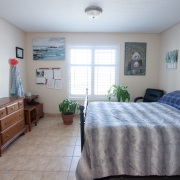 Tidy bedroom has a large window and beige painted walls. A bed with a grey and white striped bedspread is situated on the right hand side of the room against the walll. Across from the bed is a brown wooden dresser, and an end table closer to the back corner. 2 green plants are near the window at the back of the room and a black arm chair is set in the right hand corner. A picture of a Panda hangs to the right of the window, and a water landscape peice of art and message board are hanging on the wall to the left.