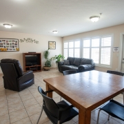 Living area with square brown table and three black chairs at the front. At the back of the room there is a black couch up against a wall of windows on the right hand side and a black lazy boy chair on the left. A brown television stand is situated against the wall at the back of the room and has a tall green plant beside it on the right. The back wall has 2 wall hangings for decoration.