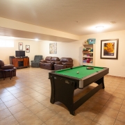 Large open and bright windowed basement room. There is a pool table set up at one end with lots of space around it. 2 brown couches and 2 lazy boy chairs are set up against the wall close to a brown tv stand and television at the other end. A shelving unit near the pool table is stocked with various items and there is a colourful painting hanging on the wall behind the pool table. 3 pictures are hanging in the tv area, one over the couch and 2 on the wall beside the tv.