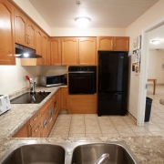 Kitchen with light brown wood cabinetry, black fridge, wall oven, counter cook top and microwave. Double sinks are surrounded by marbled beige countertops.