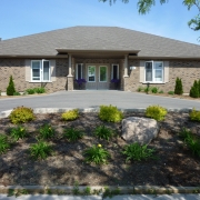 Brown brick bungalow with large double windowed entrance doors. Covered large entrance way is supported by brick and beige painted wooden pillars. There are big windows to the left and right of the house, and a large paved driveway leads to entrance. On either side of the house and in front of the driveway there are landscaped shrubs, green plants and rocks.