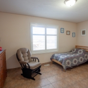 Client's bedroom has a white window and beige painted walls. A single bed with a geometric design on the bedspread is situated in the back right hand corner. A brown wooden dresser is against the wall on the left, and a comfy looking beige recliner rocker is set up near the window. 4 small works of art are hanging on the walls by the bed.