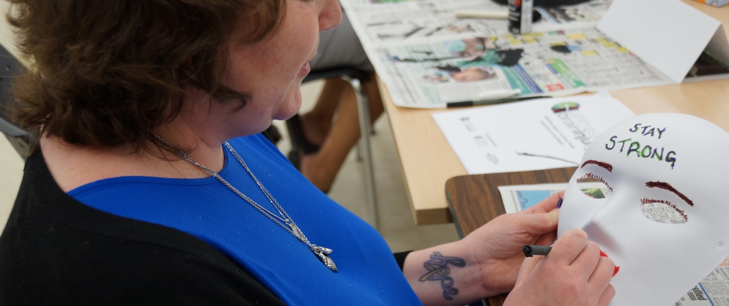 Female client sitting at a table, holding a white mask that says 'STAY STRONG' on the forehead; she is designing the mask as part of a creative expression workshop with the intention of reflecting on her personal brain injury journey.