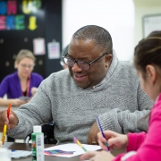 A male client and a female are sitting together at a table while they work on creative painting projects. A female staff member is sitting at another table in the room in the background.