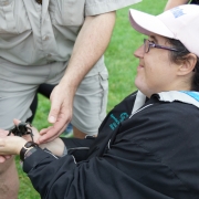 Female client sitting down with outreached hands is smiling and looking up at an exotic animal staff member while he helps to place a big black spider in the palms of her hands.