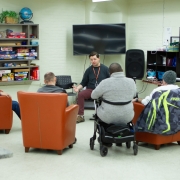 Male resource nurse sits in front of 4 male clients facilitating a presentation on health. 3 clients are sitting in big orange easy chairs, and one is sitting in front of the nurse in a wheelchair.