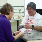 Female HIRO staff and male client sit across the table from one another while playing a game of cards. They are both holding their hand of cards towards themselves deciding what card to play next at a table.