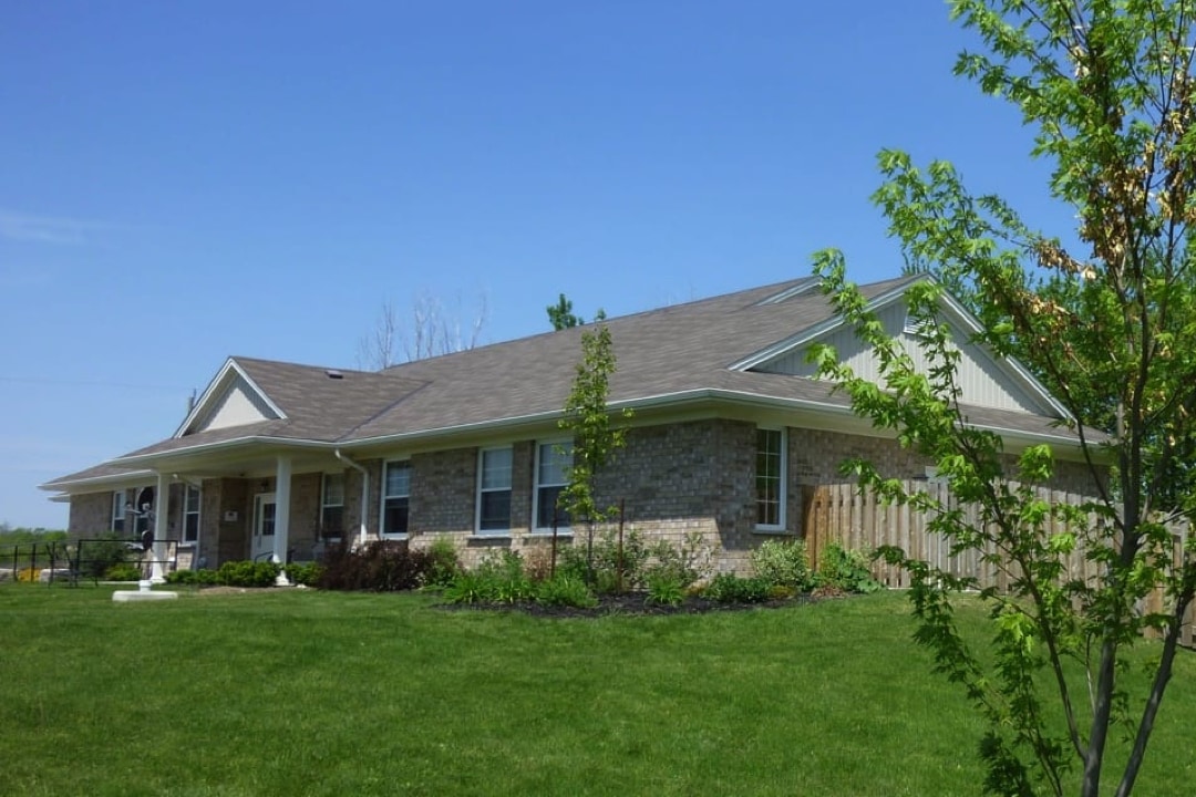 Large beige brick ranch-style bungalow has lots of front windows a covered entrance and a fenced area on the right side. It is surrounded at the front by a well maintained lush green lawn and landscaped with trees and shrubs.