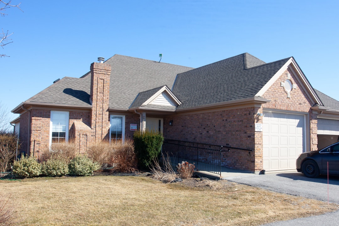 Brown brick bungalow with double car garage; accessible ramp on the left side leading up to the front door; well-maintained front lawn with trees and bushes