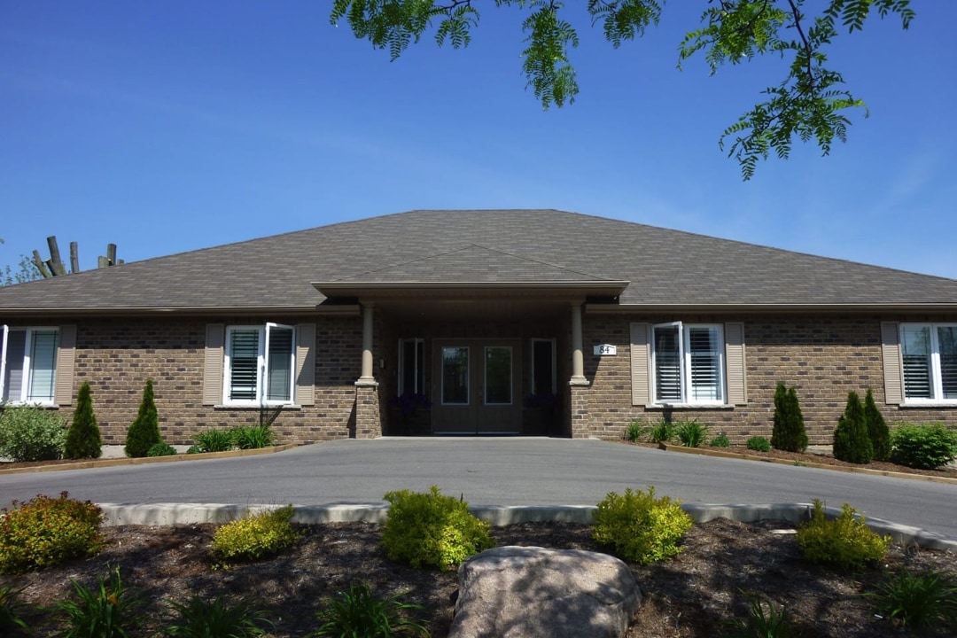 Brown brick bungalow has big front facing windows. Leading to the large covered house entrance way is a wide paved driveway. Out front of the house on either side of the entrance is green shrubery. In front of the driveway is a landscaped area with large rocks and green plants.