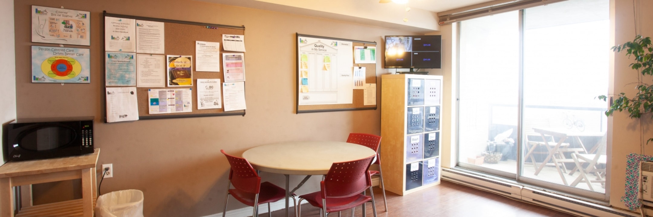 Bright room with beige painted walls has a round table and three red chairs against the wall in the middle. A black microwave sits on a wooden stand on the left hand side, and three bulletin boards with organized notices attached are hung spaced out along the wall. A filing unit has a tv screen sitting on top in the corner of the room near large window sliding doors that lead to an outdoor patio.