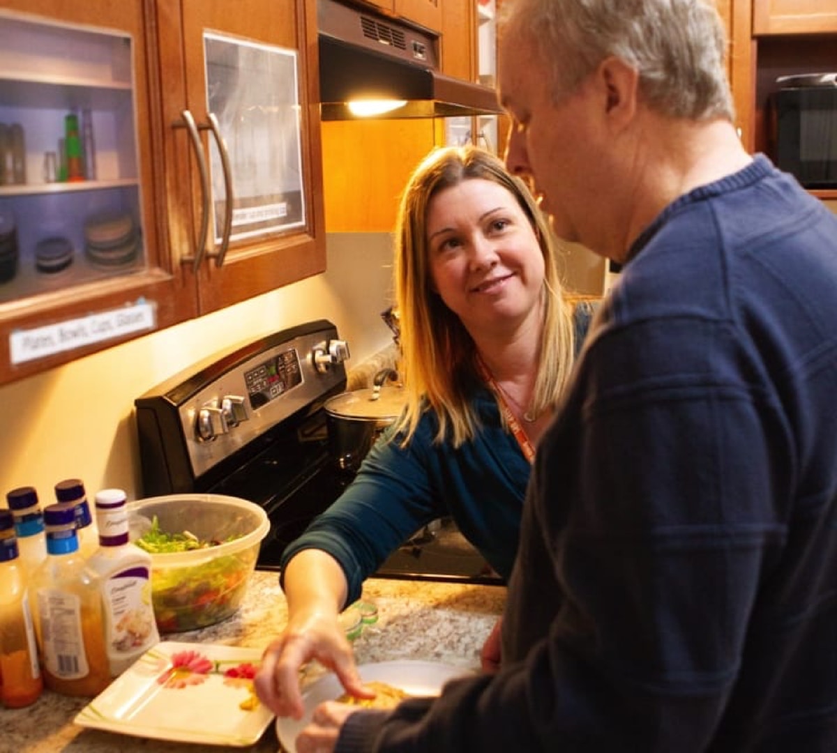 Male client is with smiling female staff member standing together at the kitchen counter preparing plates for a meal.