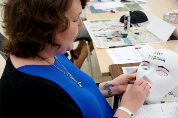 Female client sitting at a table, holding a white mask that says 'STAY STRONG' on the forehead; she is designing the mask as part of a creative expression workshop with the intention of reflecting on her personal brain injury journey.