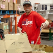 Male client is standing at a table in a large wearhouse. He is holding a cardboard box and packing tape while he works to break down the box. He is working at a food bank during a volunteer placement.