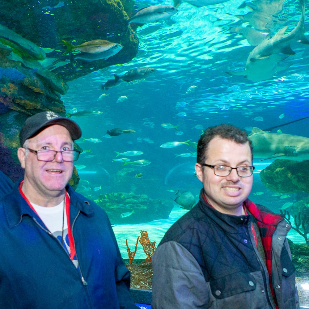 2 male clients are smiling and standing side by side in front of a huge aquarium filled with aqua blue/green water; a large green rock structure on the left; and lots of sharks swimming around in the background. They are visiting Ripley's Aquarium in Toronto during an annual client field trip.